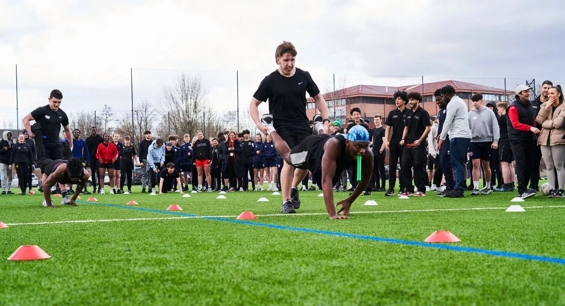 A group of learners participating in outdoor physical activities on a sports field at The Manchester College, watched by other students gathered around the area.