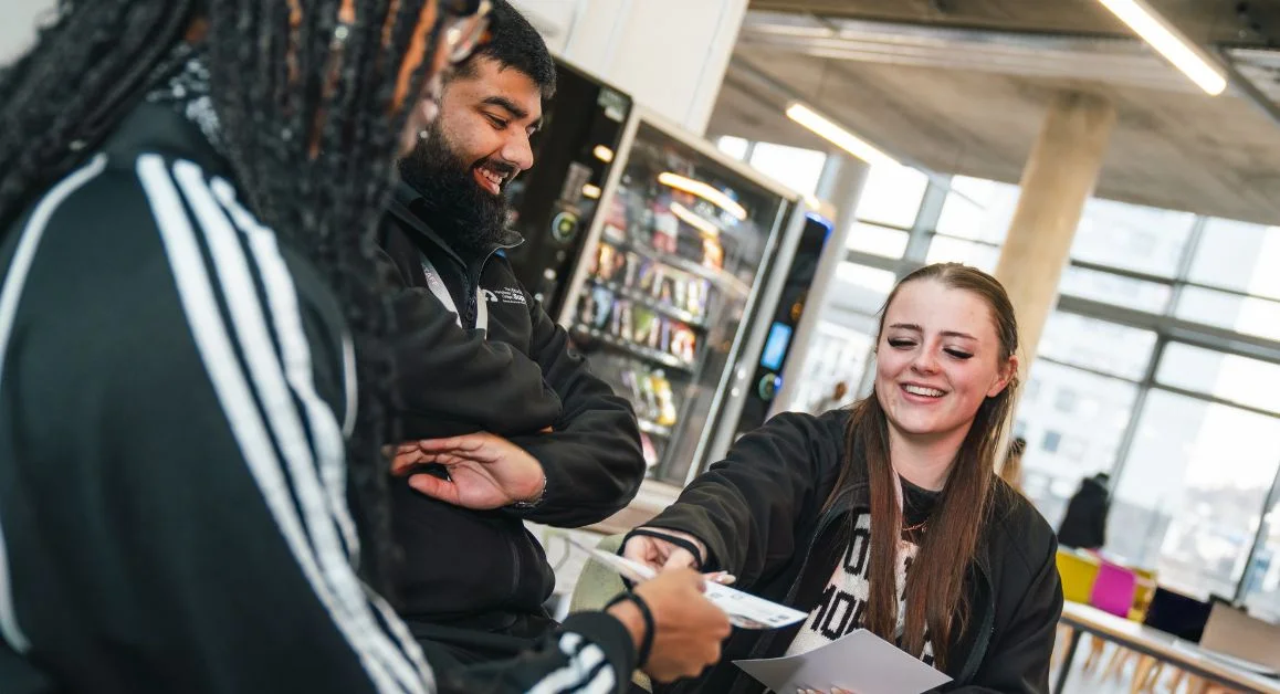Learners sitting in a college café area at The Manchester College while speaking with a staff member, with vending machines and tables in the background.
