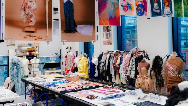 A fashion studio filled with garments on rails, mannequins, textiles, and student design work displayed across tables and hanging prints.