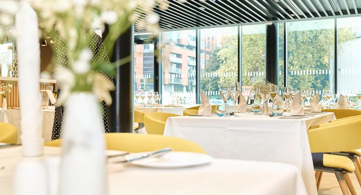 A bright, modern training restaurant at The Manchester College set with white tablecloths, folded napkins, and glassware, overlooking large windows with views of the city.