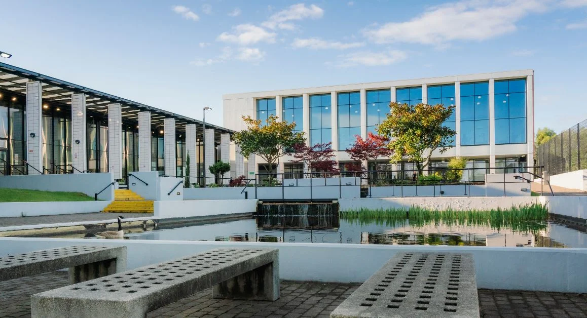 Exterior of The Manchester College Openshaw Campus, showing modern buildings with large glass windows beside a landscaped courtyard and pond.