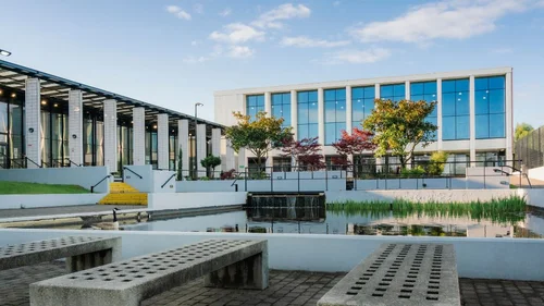 Exterior of The Manchester College Openshaw Campus, showing modern buildings with large glass windows beside a landscaped courtyard and pond.