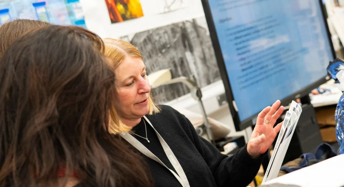 A person presenting information to others in a classroom, with a large screen displaying text in the background.