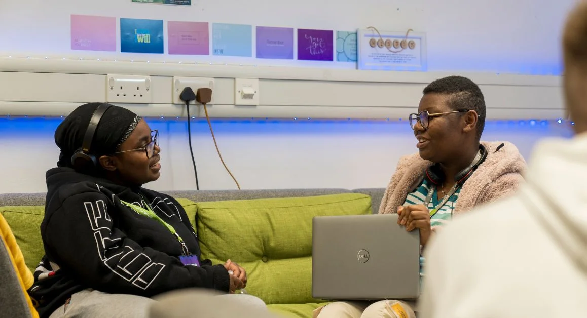 Two people sitting on a sofa in The Manchester College, talking while one holds a laptop. Colourful posters are displayed on the wall behind them.