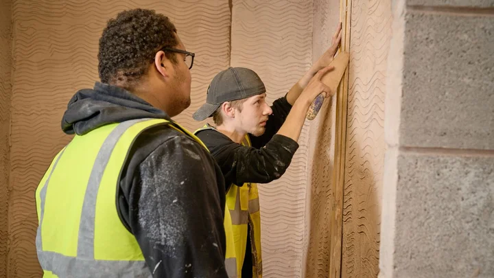 Two learners wearing high‑visibility vests working on a plastering task, applying and smoothing plaster onto an interior wall in a construction training environment.