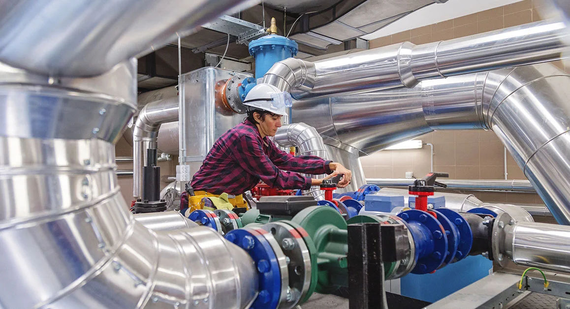 Worker operating industrial valves among large metal pipes.