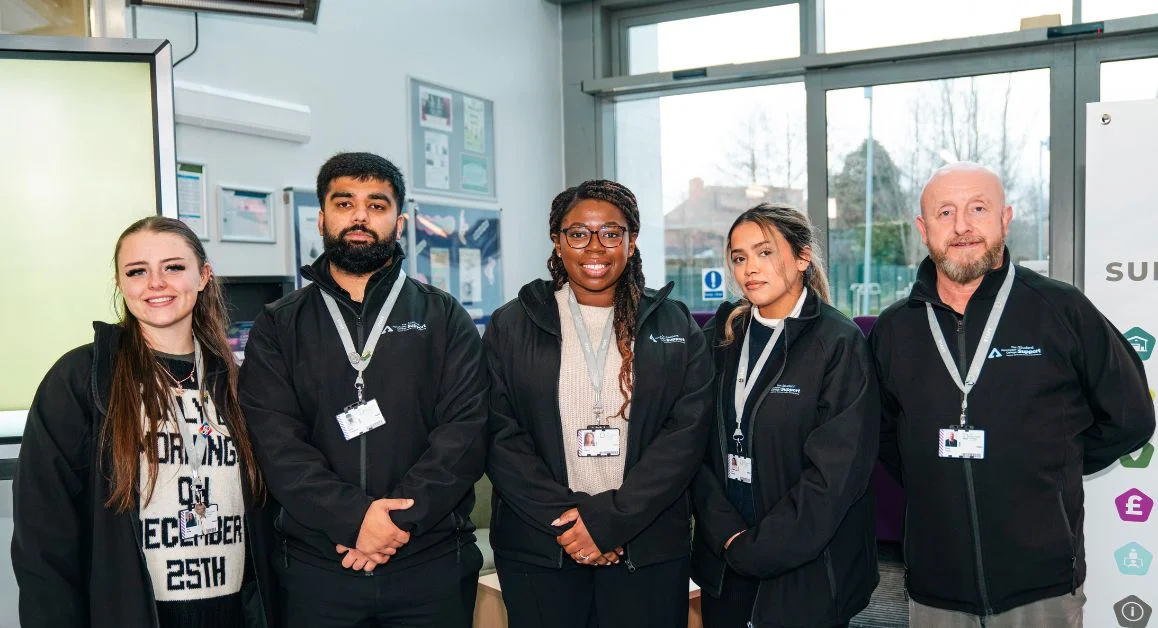 A small support team posing together in a reception area at The Manchester College, wearing matching jackets and lanyards.
