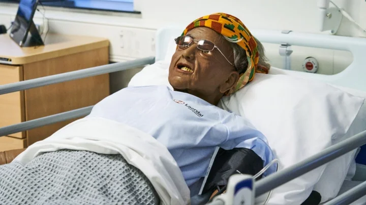 Patient manikin lying in a hospital bed during a clinical skills training session, with medical equipment positioned at the bedside.
