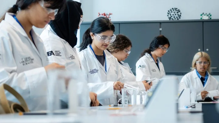 Science learners in lab coats carrying out a practical experiment using measuring cylinders and equipment on a laboratory bench.