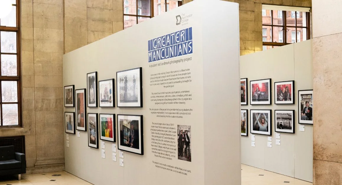 An art exhibition display inside a large public building, featuring framed photographs arranged on white panels under the title “Greater Mancunians.”