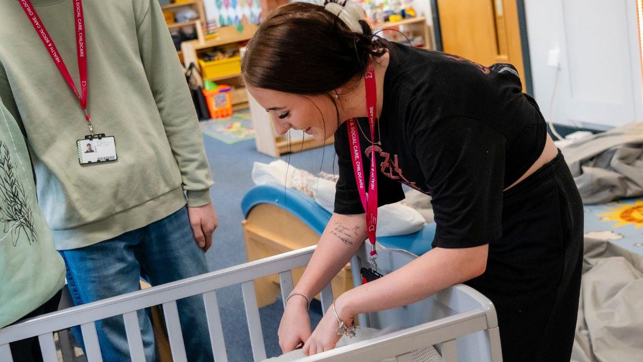 Learner placing doll into a cot during a practical childcare session in an early years classroom at The Manchester College, with training equipment and learning resources visible in the background.