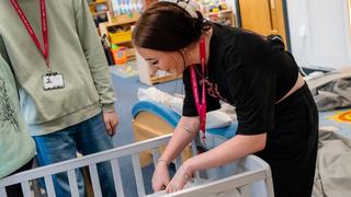 Learner placing doll into a cot during a practical childcare session in an early years classroom at The Manchester College, with training equipment and learning resources visible in the background.