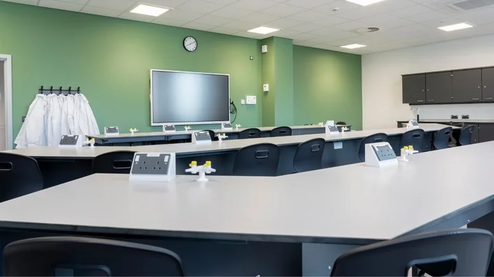 A modern science laboratory with long bench tables, gas taps, lab stools, an interactive screen, and lab coats hanging on the wall.