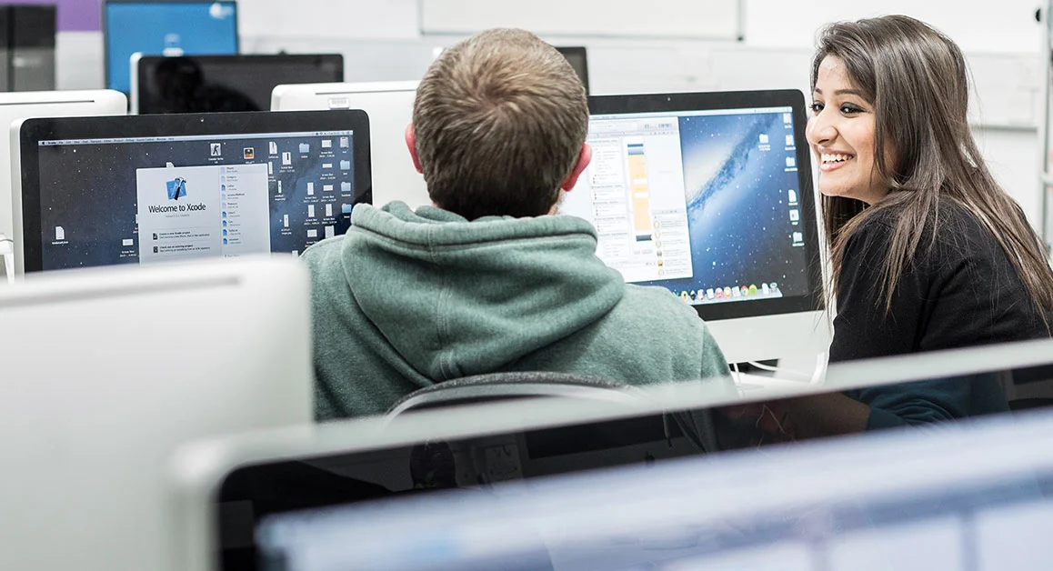 Two people collaborating at desktop computers in a computer lab.