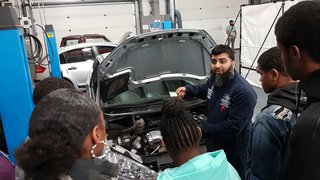 A group of students listen closely to a mechanic explaining about a car engine.