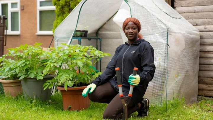 A learner kneeling in a garden area holding hedge clippers next to a greenhouse and potted plants.