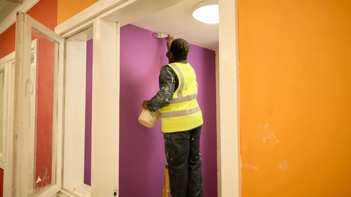 A learner wearing a high‑visibility vest painting a purple interior wall using a roller inside a brightly coloured training environment.