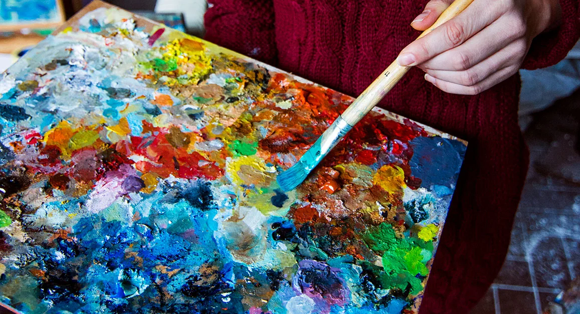 Person dipping a paintbrush into a colourful artist’s palette.