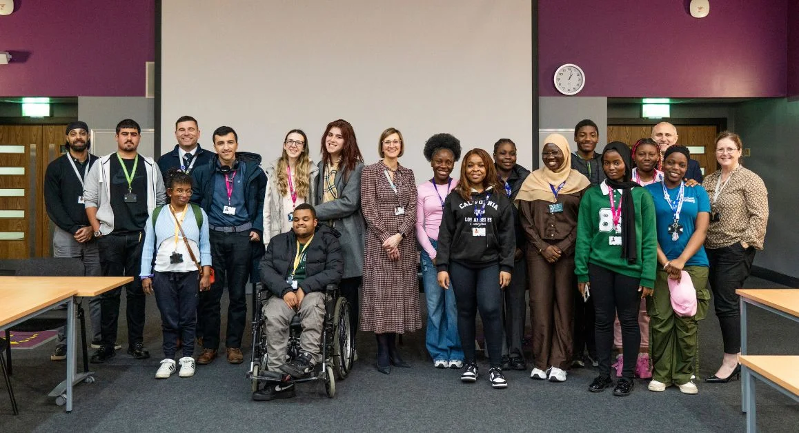 A large group of learners and the principal of The Manchester College standing together in a classroom space, all wearing lanyards, with a projector screen in the background.