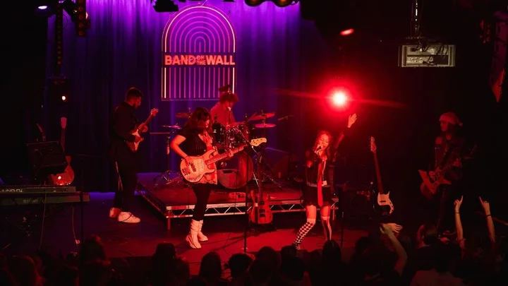 A live music performance on stage at Band on the Wall, featuring a full band with guitars, drums, keyboards and a vocalist performing under red and purple stage lighting.