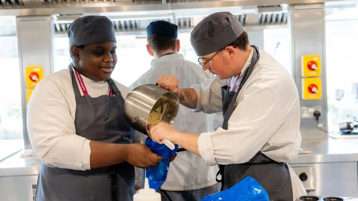 Catering learners working together in a professional training kitchen, transferring mixture from a saucepan into a piping bag.
