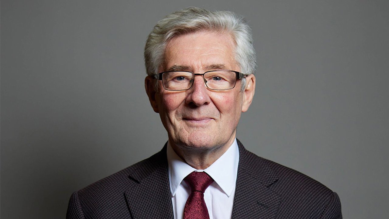 A portrait of Sir Tony Lloyd MP. He's wearing a suit with a red tie and standing in front of a grey background.