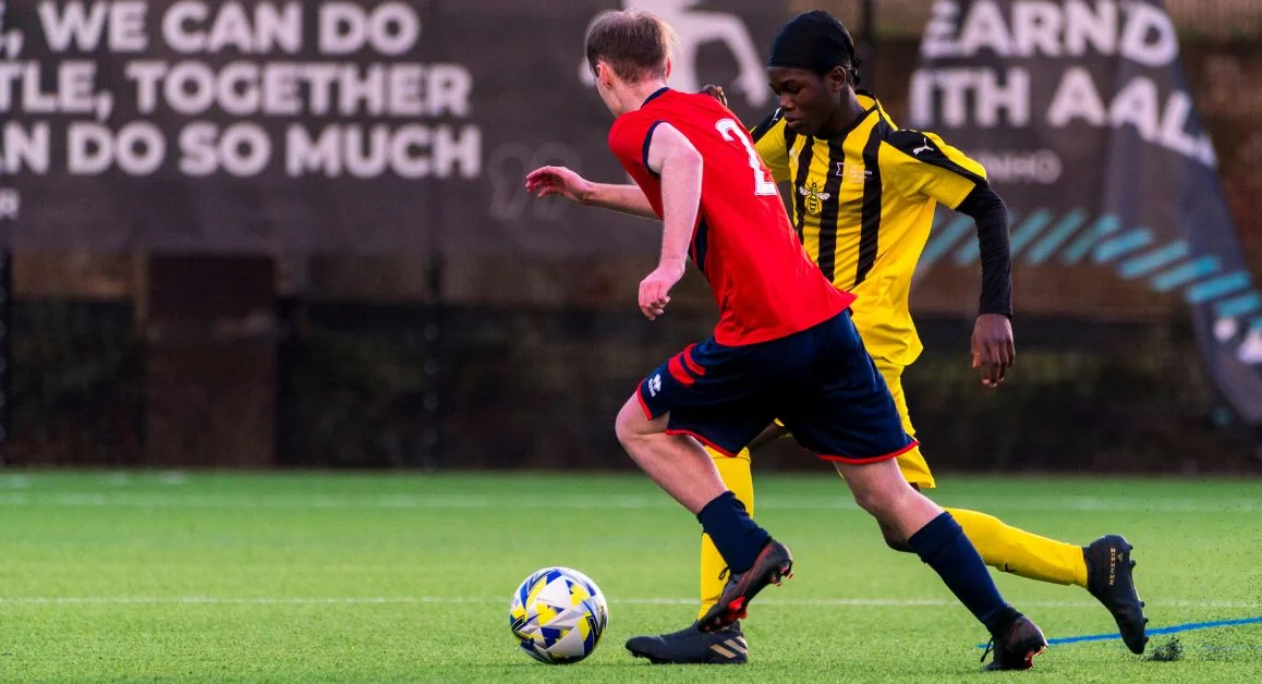 Two football players in action on an outdoor pitch, one running with the ball while another defends closely.