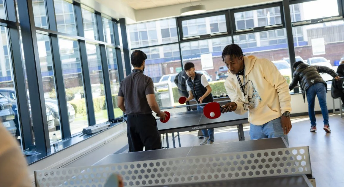 Students playing table tennis in a bright social space at The Manchester College with floor‑to‑ceiling windows and outdoor views.