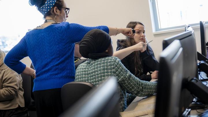 A TMC tutor supports students during a classroom activity, pointing towards a computer screen while students work collaboratively at their workstations.