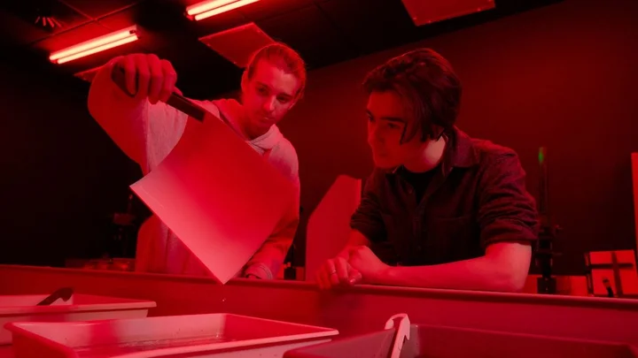 Learners developing a photograph in a darkroom, examining a print under red safelights during an analogue photography session.