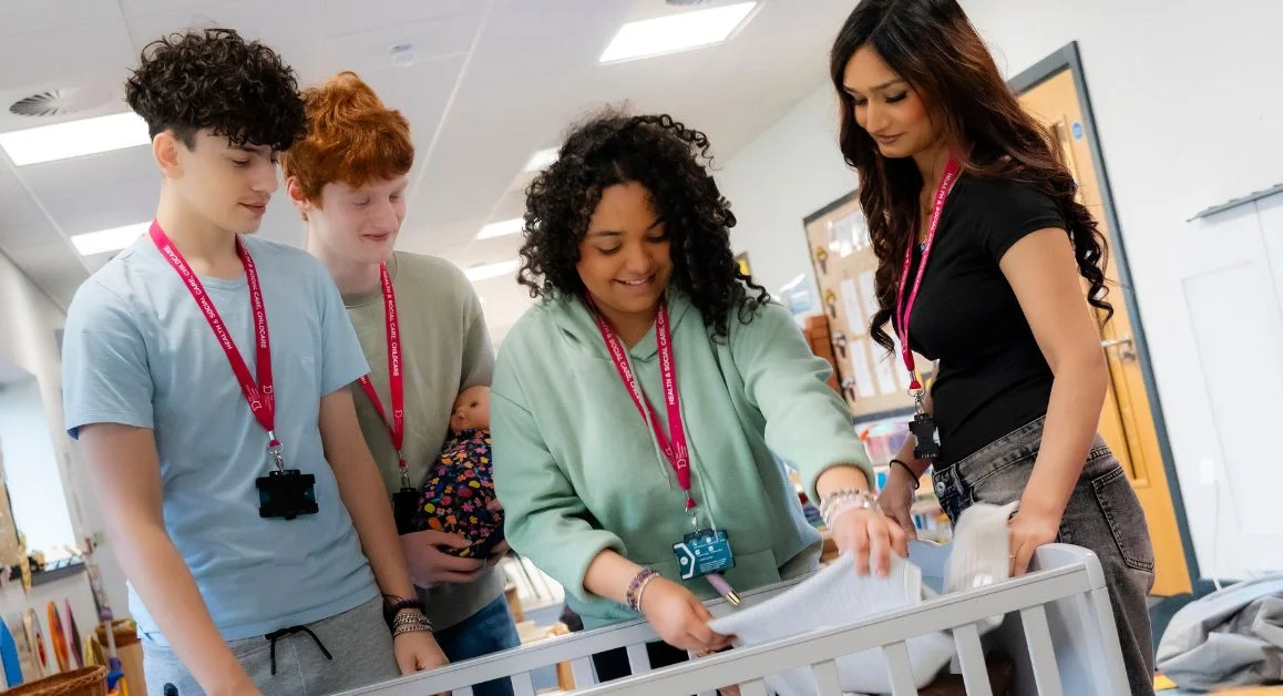 A group of learners and a teacher in a childcare classroom at The Manchester College practising how to prepare a cot, with educational toys and learning resources visible around the room.