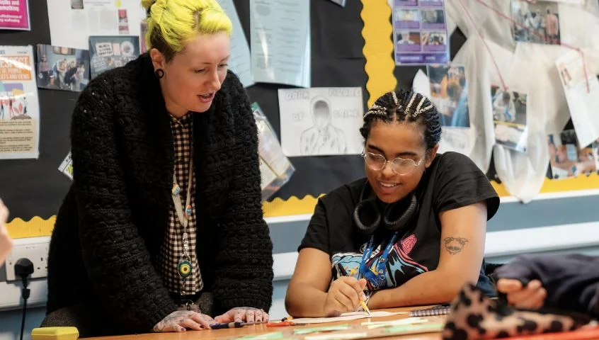 A classroom scene at The Manchester College where a staff member is supporting a learner with written work, surrounded by educational displays on the walls.