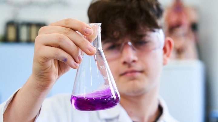 Student in a lab coat and safety goggle observing a breaker of purple liquid