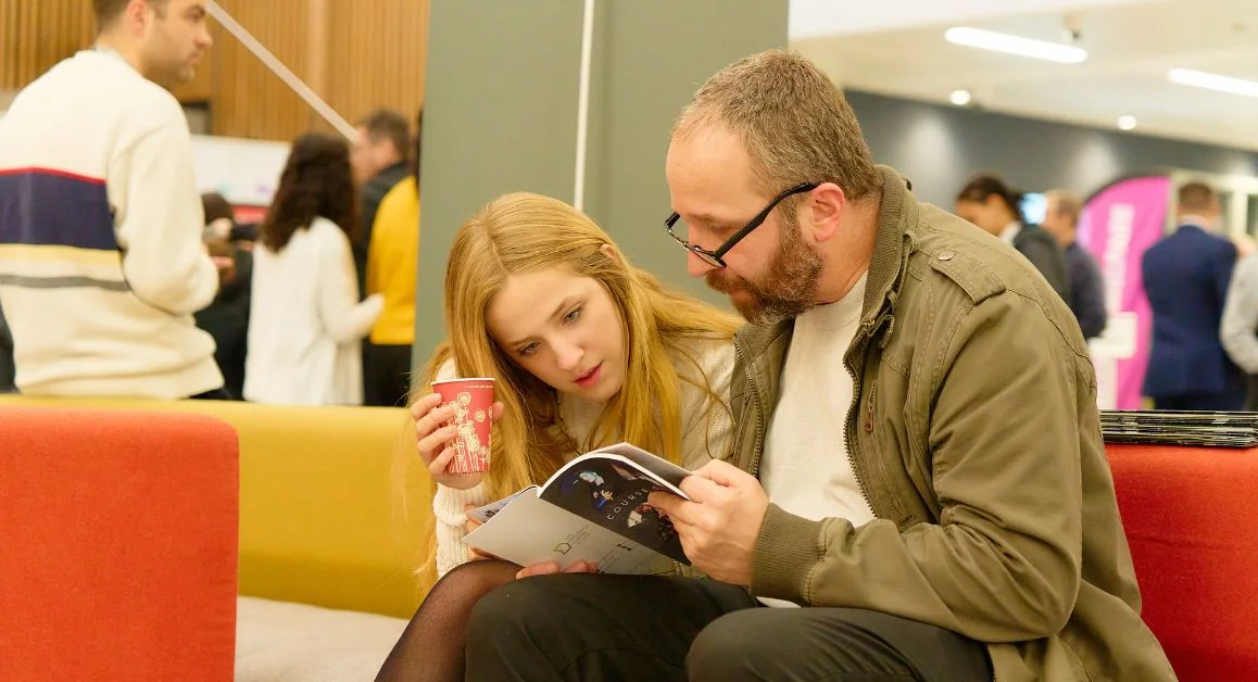 Parent and prospective student sitting on a sofa in a college common area, looking at a course guide together while others walk and talk in the background.