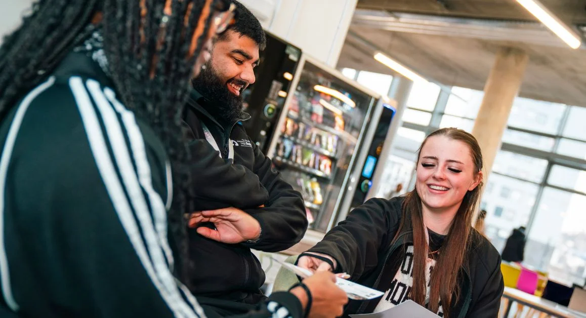 Three individuals sitting together in The Manchester College café area, discussing a leaflet, with vending machines and tables in the background.