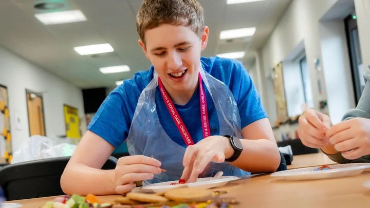 A learner in an education and early years classroom taking part in a hands‑on activity, using craft materials at a table while wearing a protective apron.