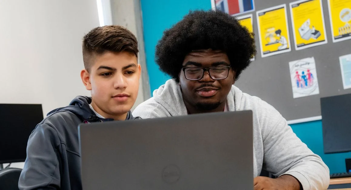 Two students seated at a desk, looking at a laptop together in a classroom with posters and computers in the background.