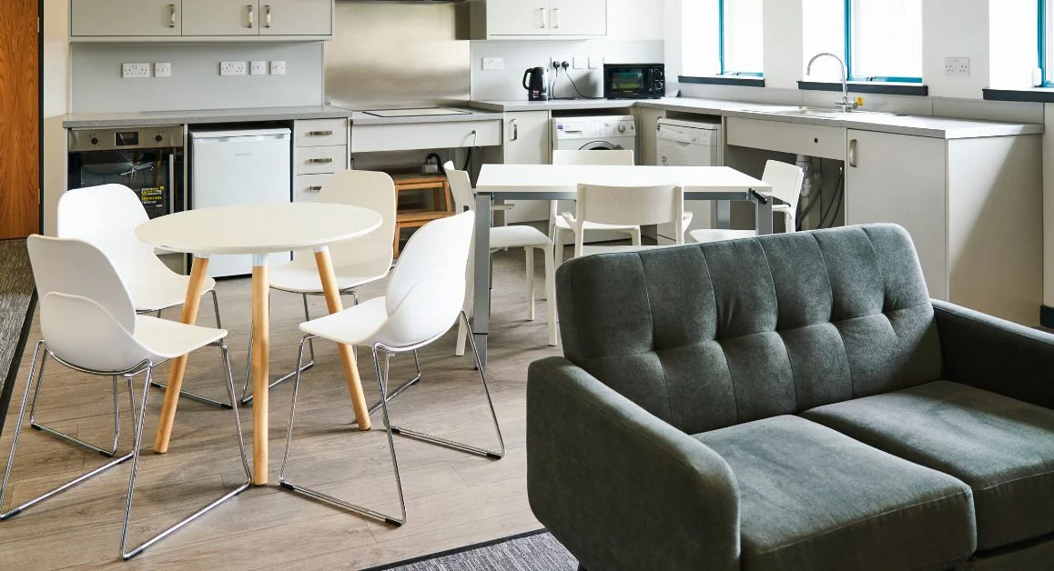 A modern student flat with shared kitchen and lounge area with white cabinets, two dining tables with chairs, and a grey sofa.