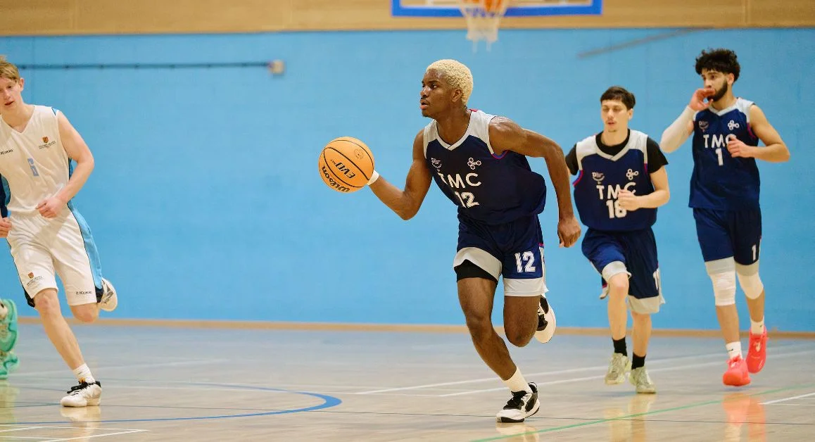Basketball players in TMC sports kit running down an indoor court, with one player dribbling the ball ahead of teammates.