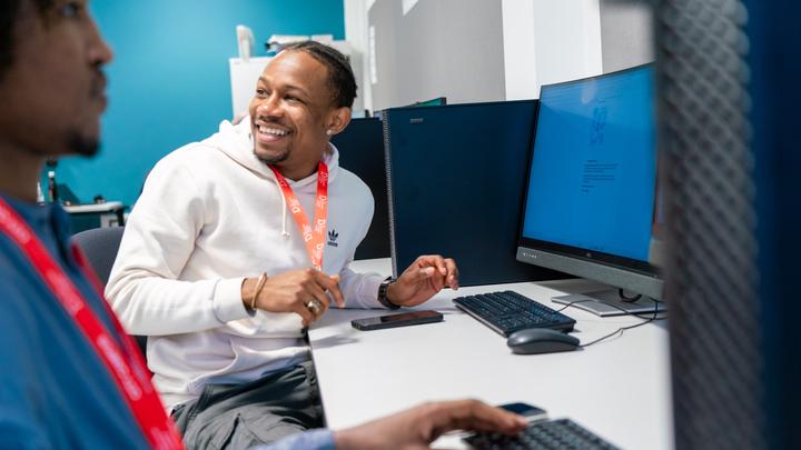 Two students seated at computer workstations in a modern classroom, discussing something on a desktop screen while working with keyboards and other equipment.