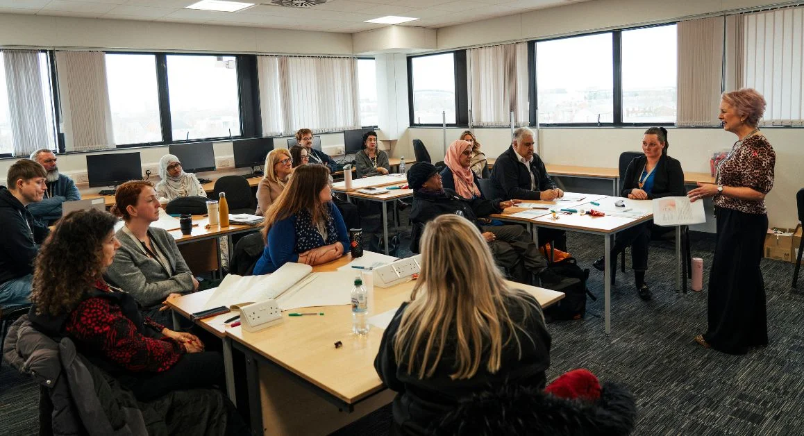 A classroom with staff members from The Manchester College seated around tables while a facilitator stands at the front presenting to the group.