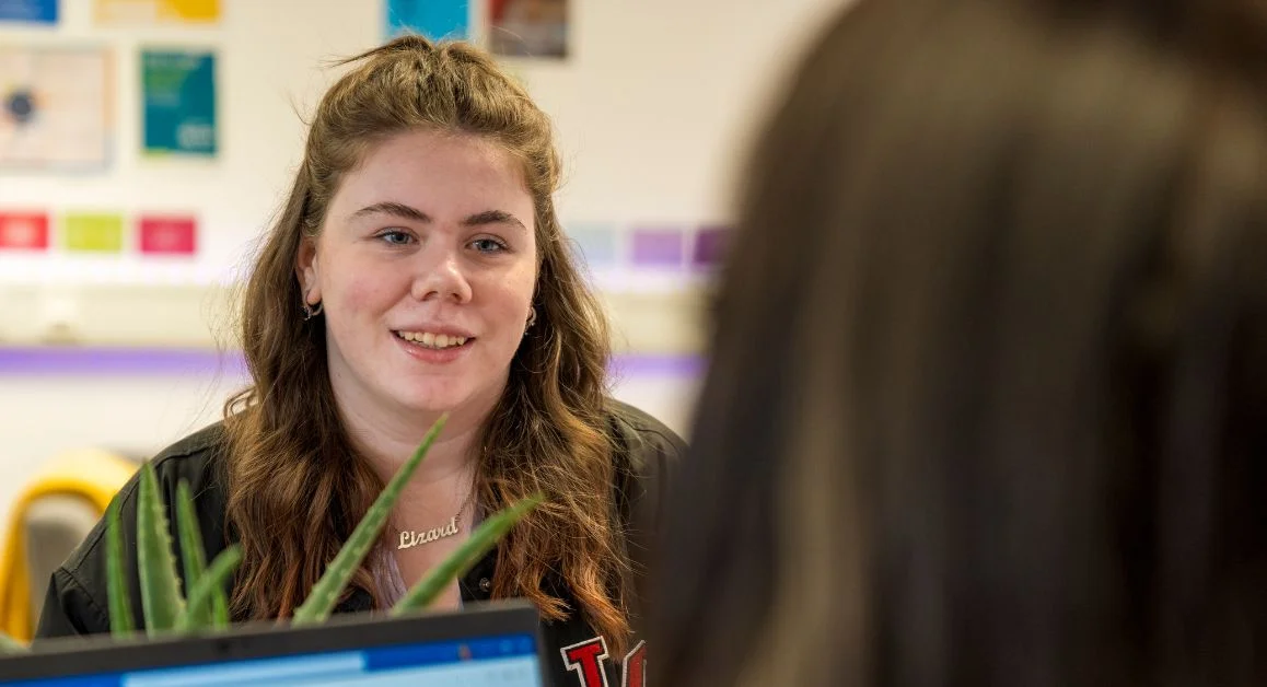 A close‑up of a student seated at a support desk in The Manchester College wellbeing area, with leaflets and posters visible on the wall.