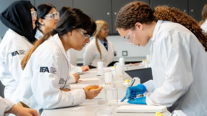 Science learners in lab coats working together at a laboratory bench, recording results and using pipettes and beakers during a practical experiment.