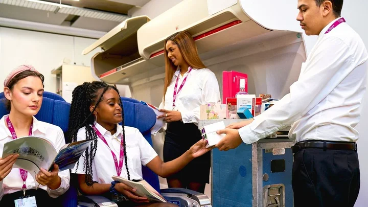 Cabin crew trainees practising in‑flight service, using a trolley to distribute items to seated learners during aviation training.