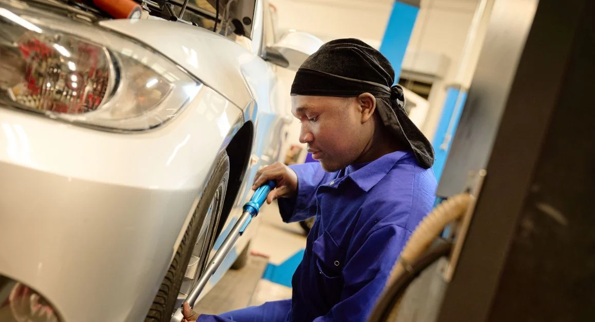 An automotive learner from The Manchester College in workshop overalls using a tool to work on the wheel area of a car in a training garage.