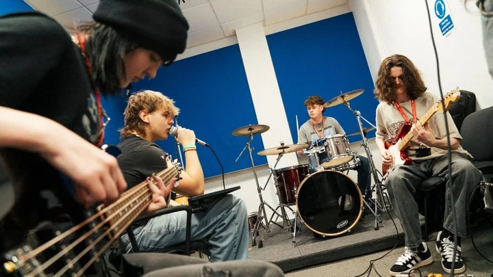 A group of music learners rehearsing together in a practice room, playing bass, electric guitar, drums, and vocals.