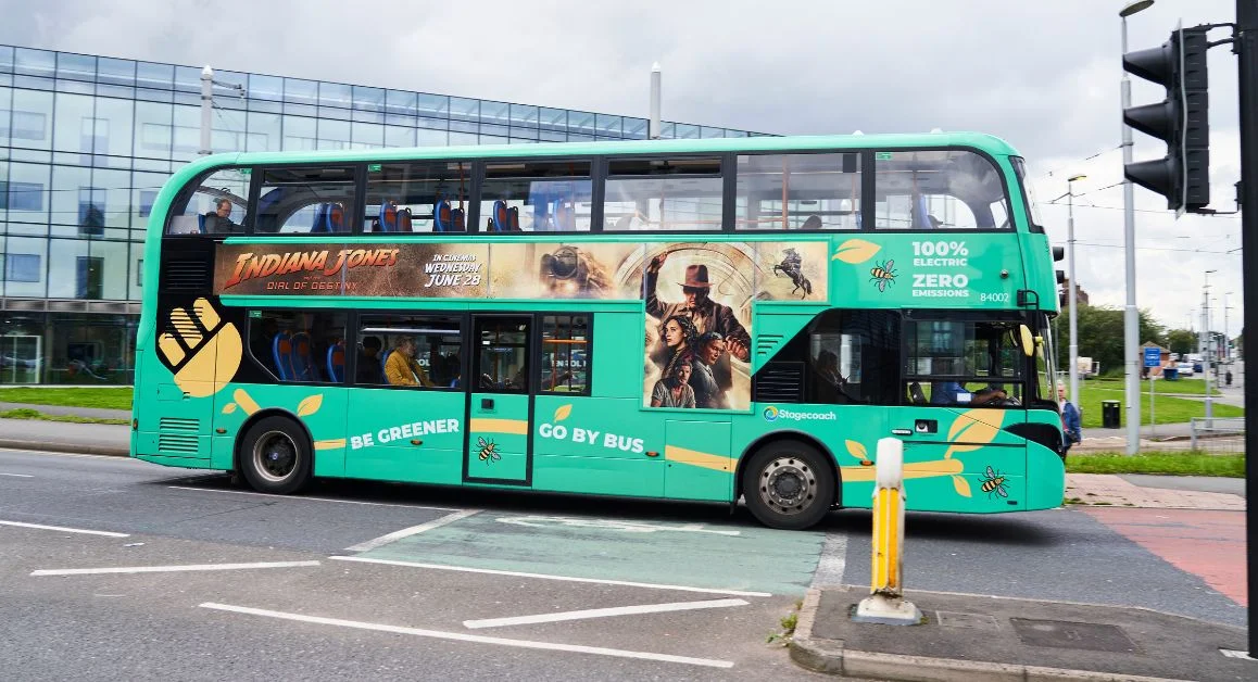 A green double‑decker bus with colourful branding and an advertisement on the side, driving through a modern city area.
