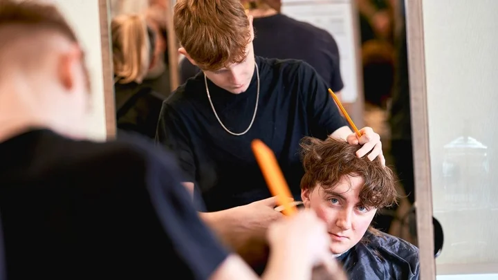 A learner practising a haircut in a barbering training session, using a comb and scissors in front of a salon mirror.